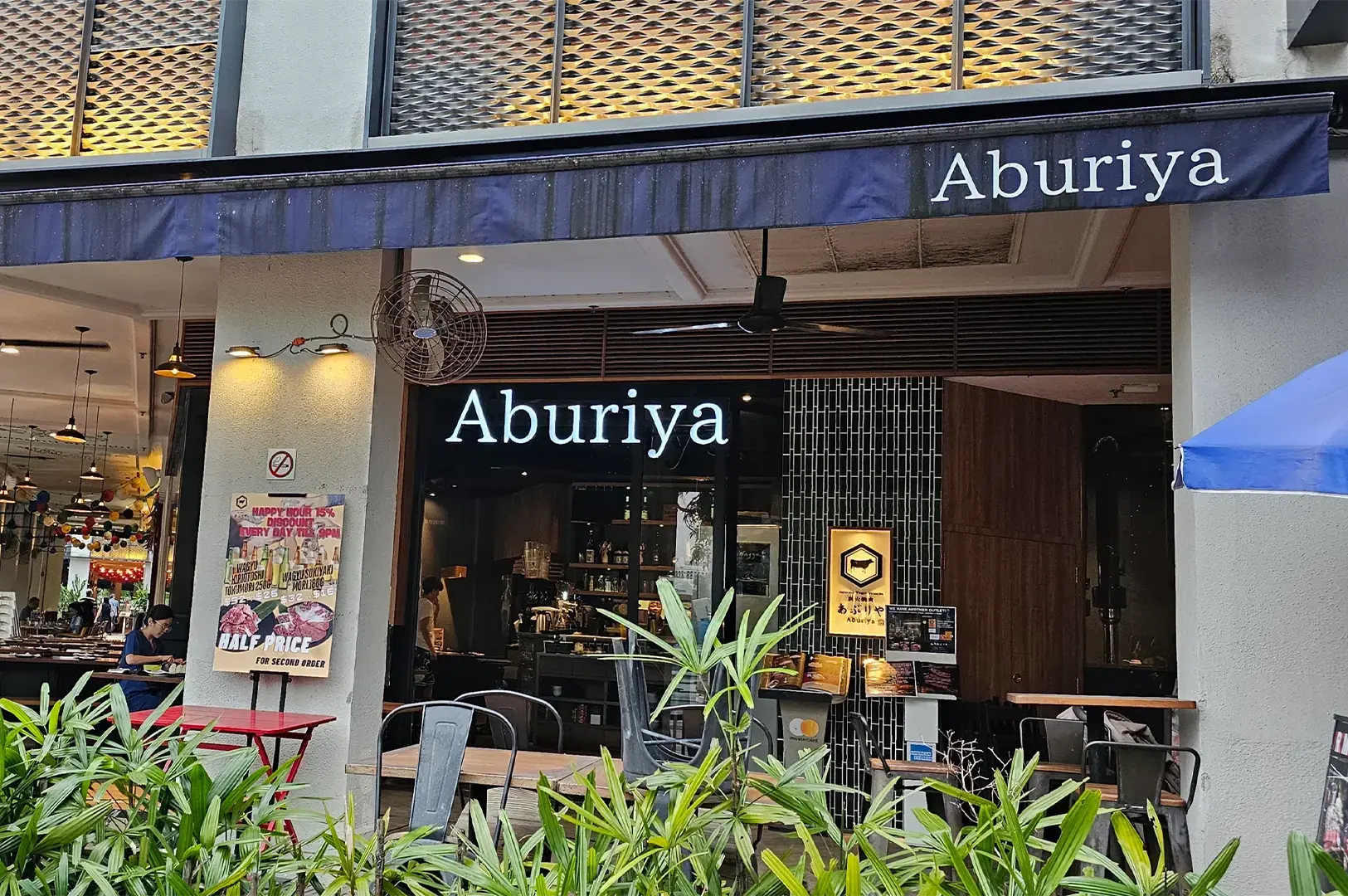 Storefront of Aburiya Japanese restaurant with an awning, outdoor dining tables, potted plants, and promotional posters.