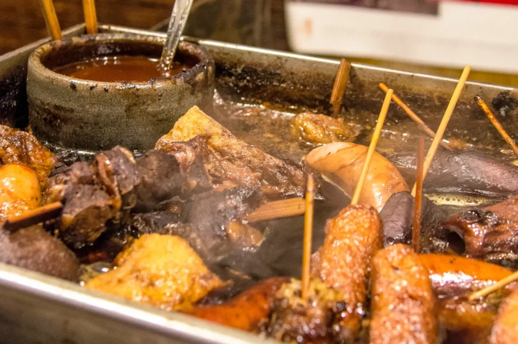 A close-up of a hot metal grill tray with sizzling fried food, including sausages and pieces of chicken or fish, with wooden skewers sticking up and oil bubbling around them.