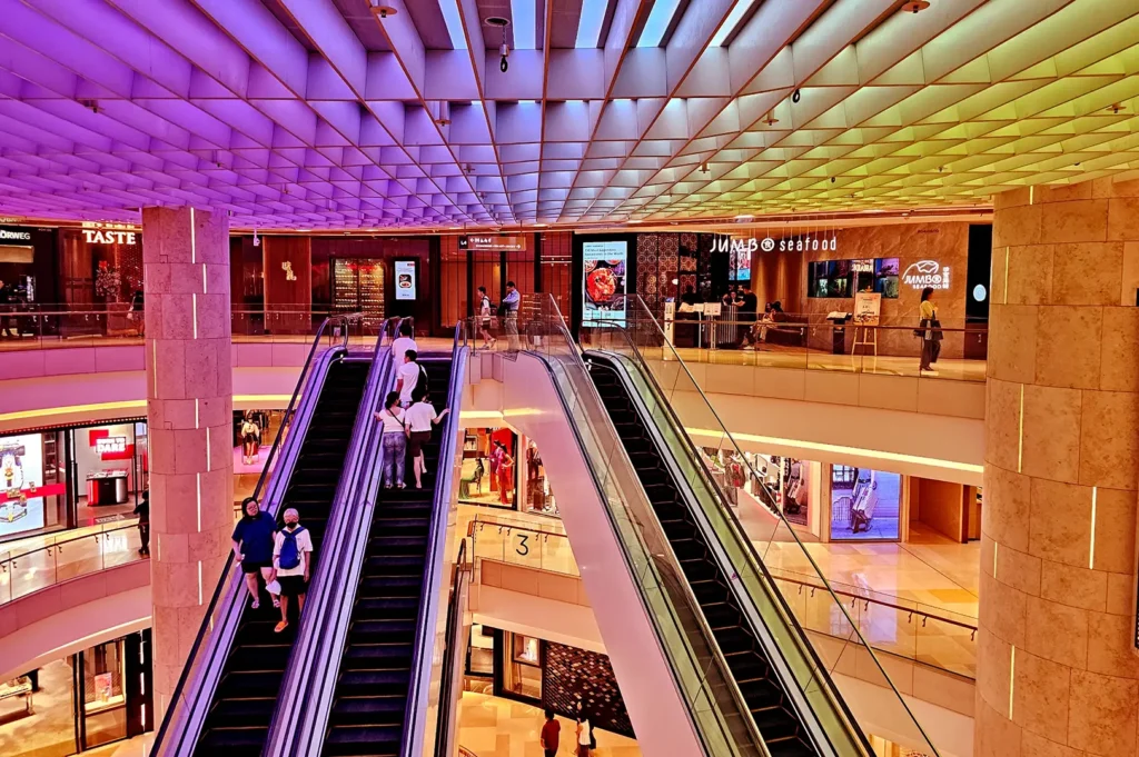 Vibrant multi-level ION Orchard interior with parallel escalators, illuminated storefronts, restaurants, shoppers, and a colourful geometric ceiling.