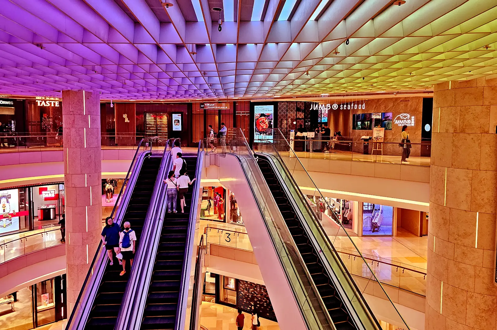 Vibrant multi-level ION Orchard interior with parallel escalators, illuminated storefronts, restaurants, shoppers, and a colourful geometric ceiling.