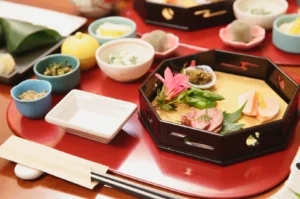 Japanese meal set on a red tray: a black lacquer bento with assorted vegetables, meat, and pink garnish, plus small side dishes in bowls and cups, chopsticks, and dipping sauces.