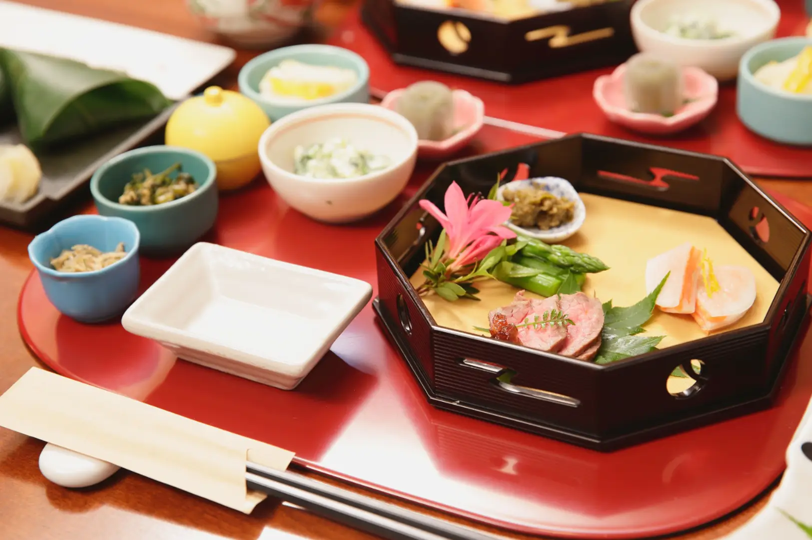 Japanese meal set on a red tray: a black lacquer bento with assorted vegetables, meat, and pink garnish, plus small side dishes in bowls and cups, chopsticks, and dipping sauces.