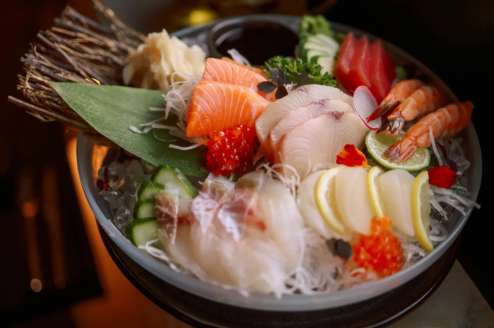 A close-up bowl of sashimi and seafood topped with salmon, red berries, cucumber, lemon slices, and herbs, with chopsticks resting beside it.
