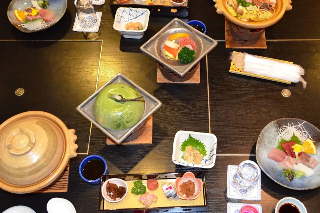 Top-down view of a Japanese-style multi-course meal on a wooden table, with small bowls and plates of colorful dishes, sushi, desserts, sauces, and drinks.