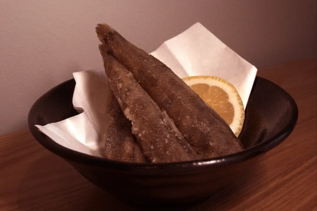 Three fried mehikari fish served in a black bowl with a lemon slice and paper liner.