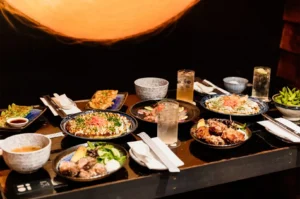 A table spread featuring various Japanese izakaya dishes, including okonomiyaki, chicken karaage, edamame, and cocktails.