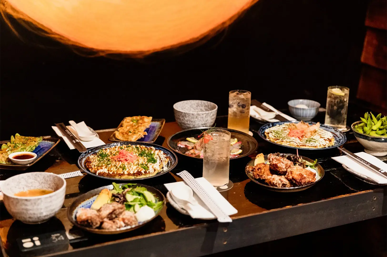 A table spread featuring various Japanese izakaya dishes, including okonomiyaki, chicken karaage, edamame, and cocktails.