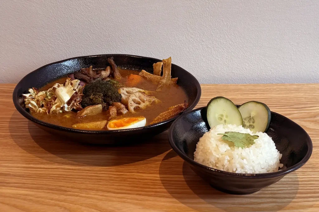 Japanese soup curry with vegetables, mushrooms, meat, and half a boiled egg served with white rice, cucumber slices, and coriander in black bowls.