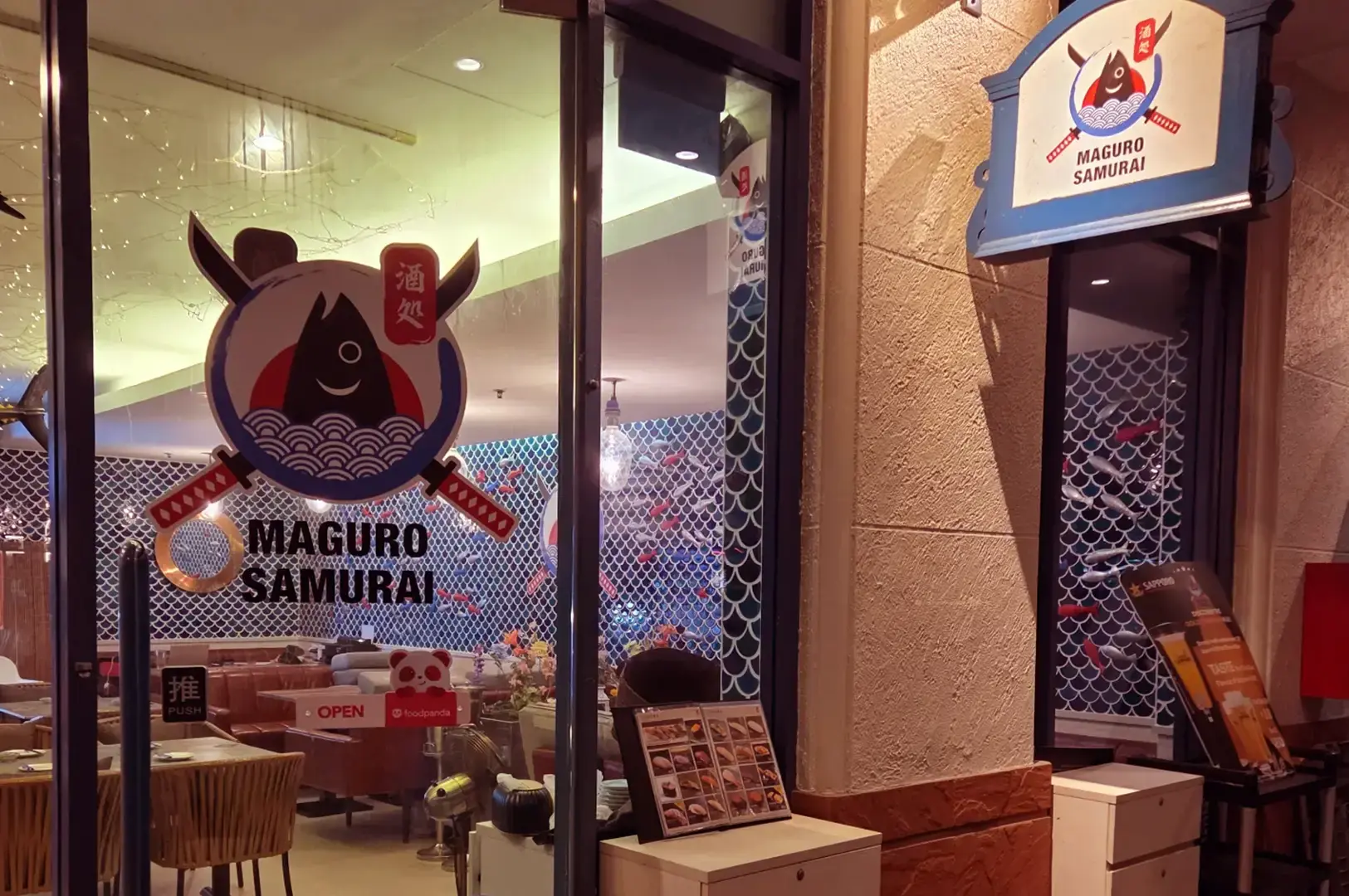 Entrance to Maguro Samurai with the fish-and-samurai logo on the glass door and exterior sign, showing the dining room inside.