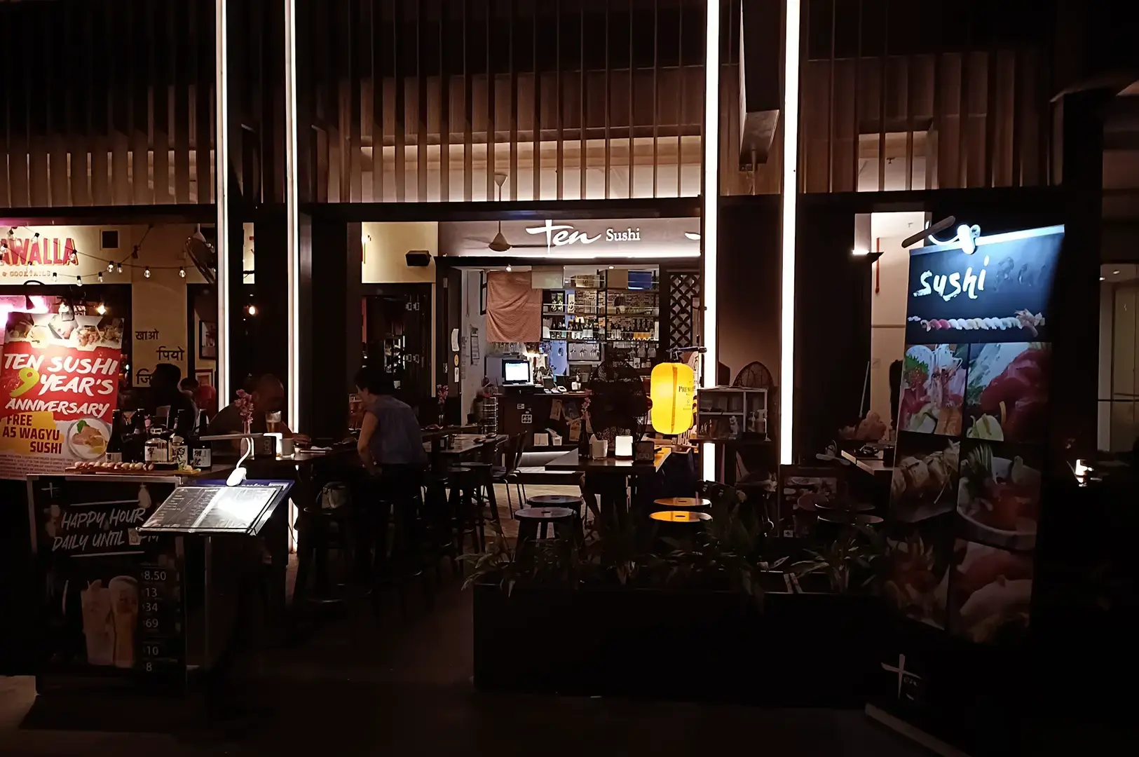 Dimly lit TEN Sushi Bar entrance with indoor tables, diners, illuminated signage, a sushi display, and a promotional board.