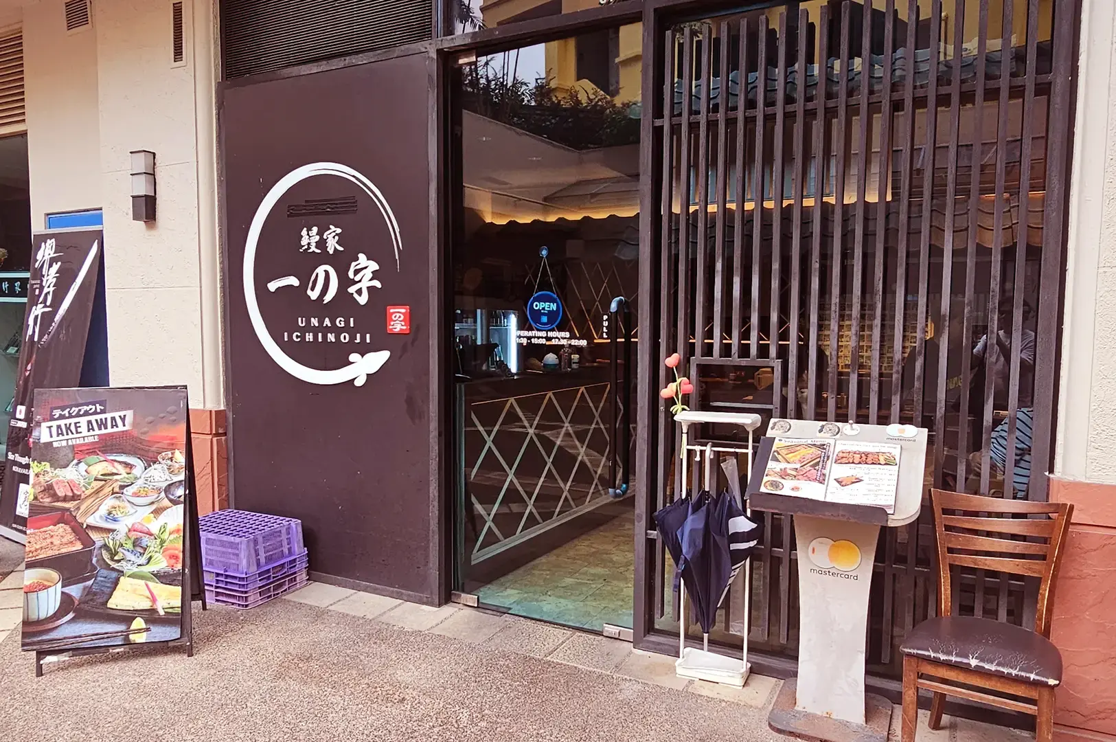 Unagiya Ichinoji entrance with its circular logo, glass doorway, takeaway sign, menu stand, umbrellas, and outdoor chair.