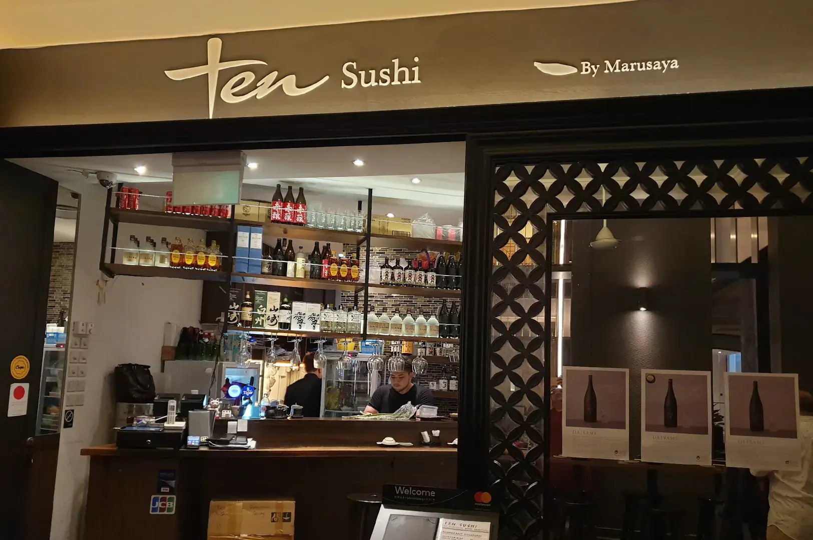 Storefront view of Ten Sushi by Marusaya showing the bar counter, liquor shelves, and staff preparing food.