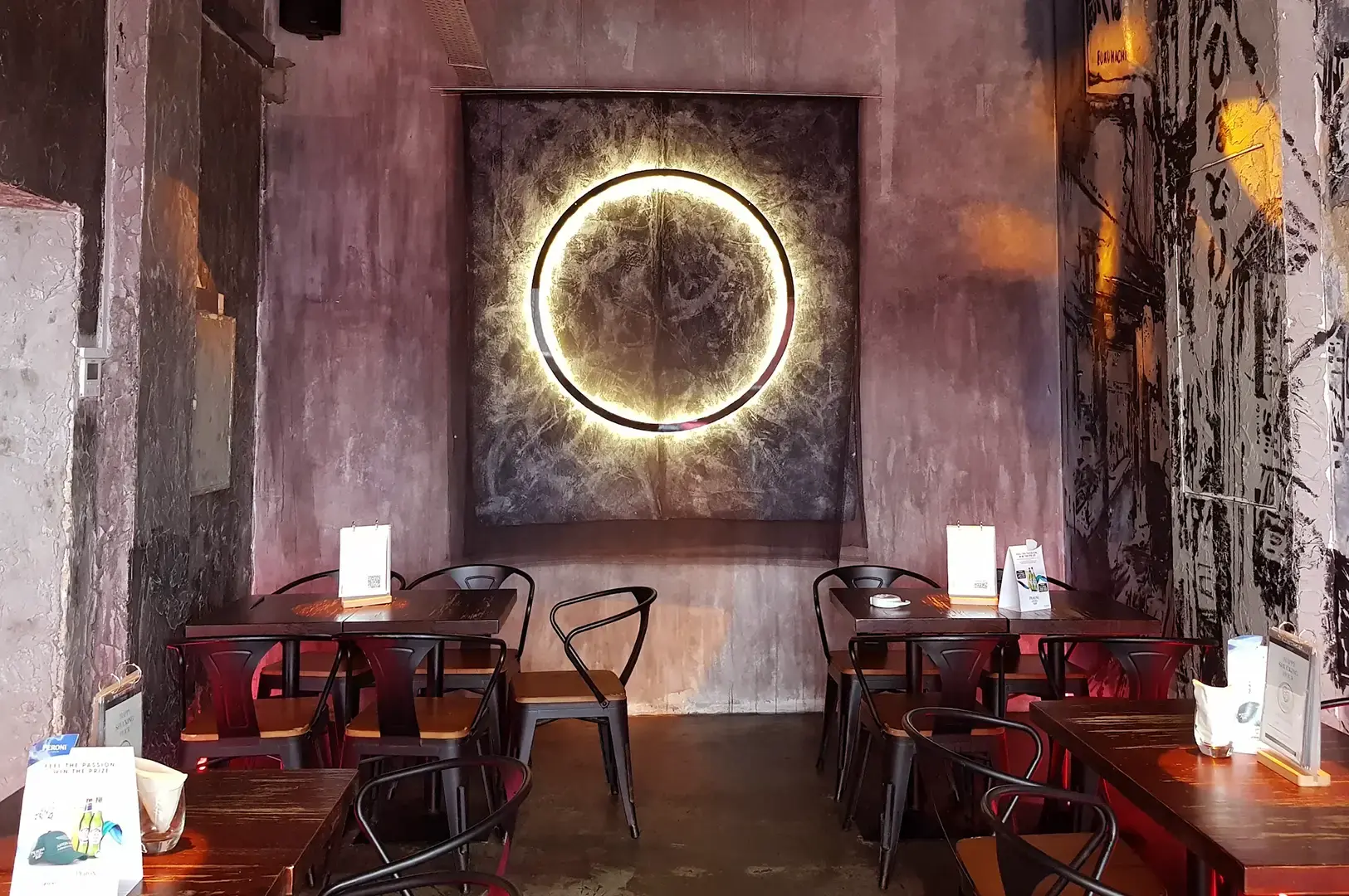 Empty TAAN IZA-BAR dining area with small tables, black chairs, textured walls, and a glowing circular wall light.