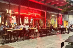 Busy Tono Izakaya entrance glowing with red lighting, with diners seated at outdoor tables along a covered shopping-centre walkway.