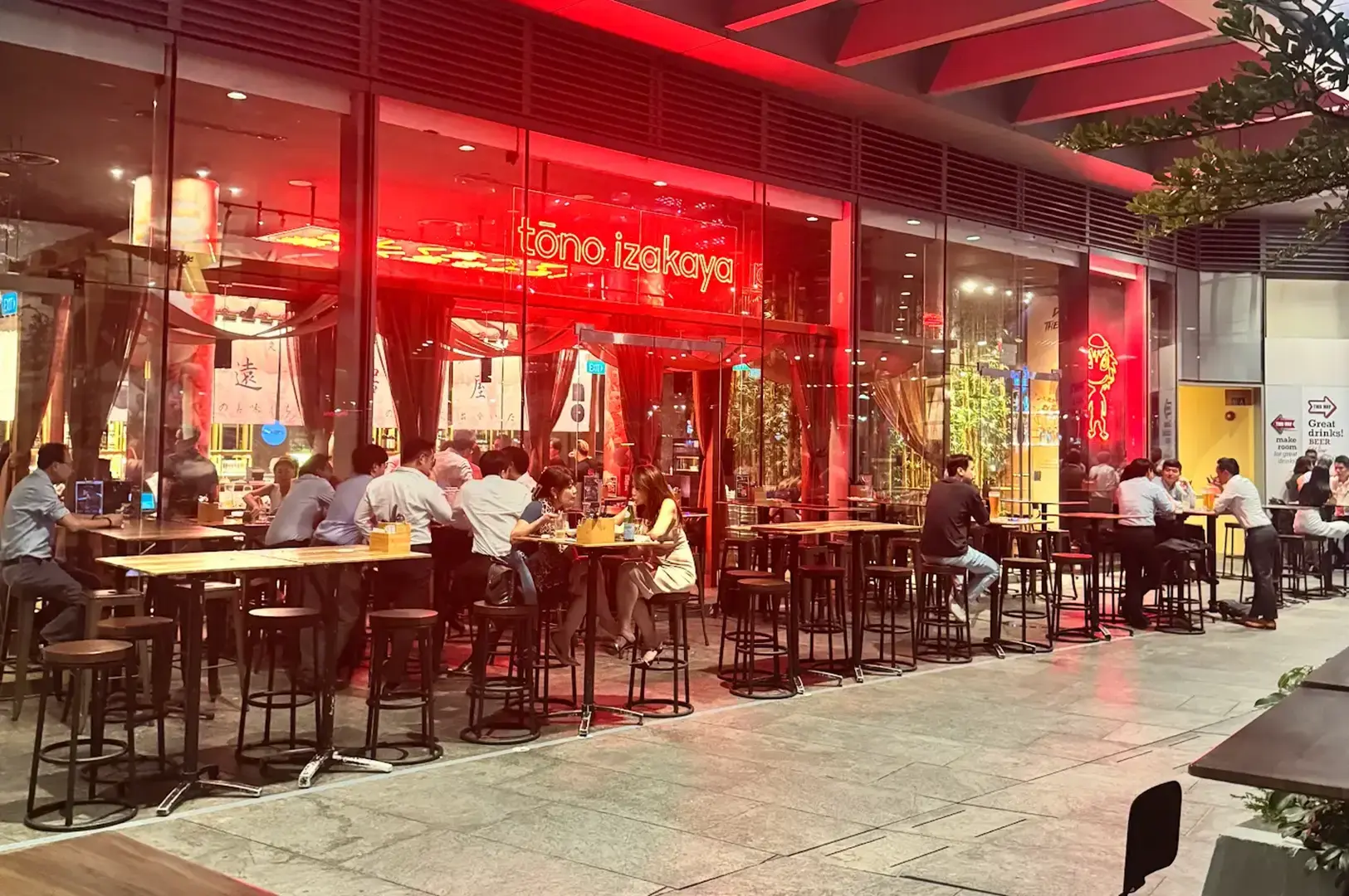 Busy Tono Izakaya entrance glowing with red lighting, with diners seated at outdoor tables along a covered shopping-centre walkway.