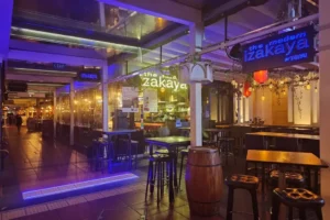 Exterior view of "The Modern Izakaya by Tatsu" featuring al fresco dining tables, high bar stools, a decorative wooden barrel, red lanterns, and a glowing blue sign under a covered walkway.